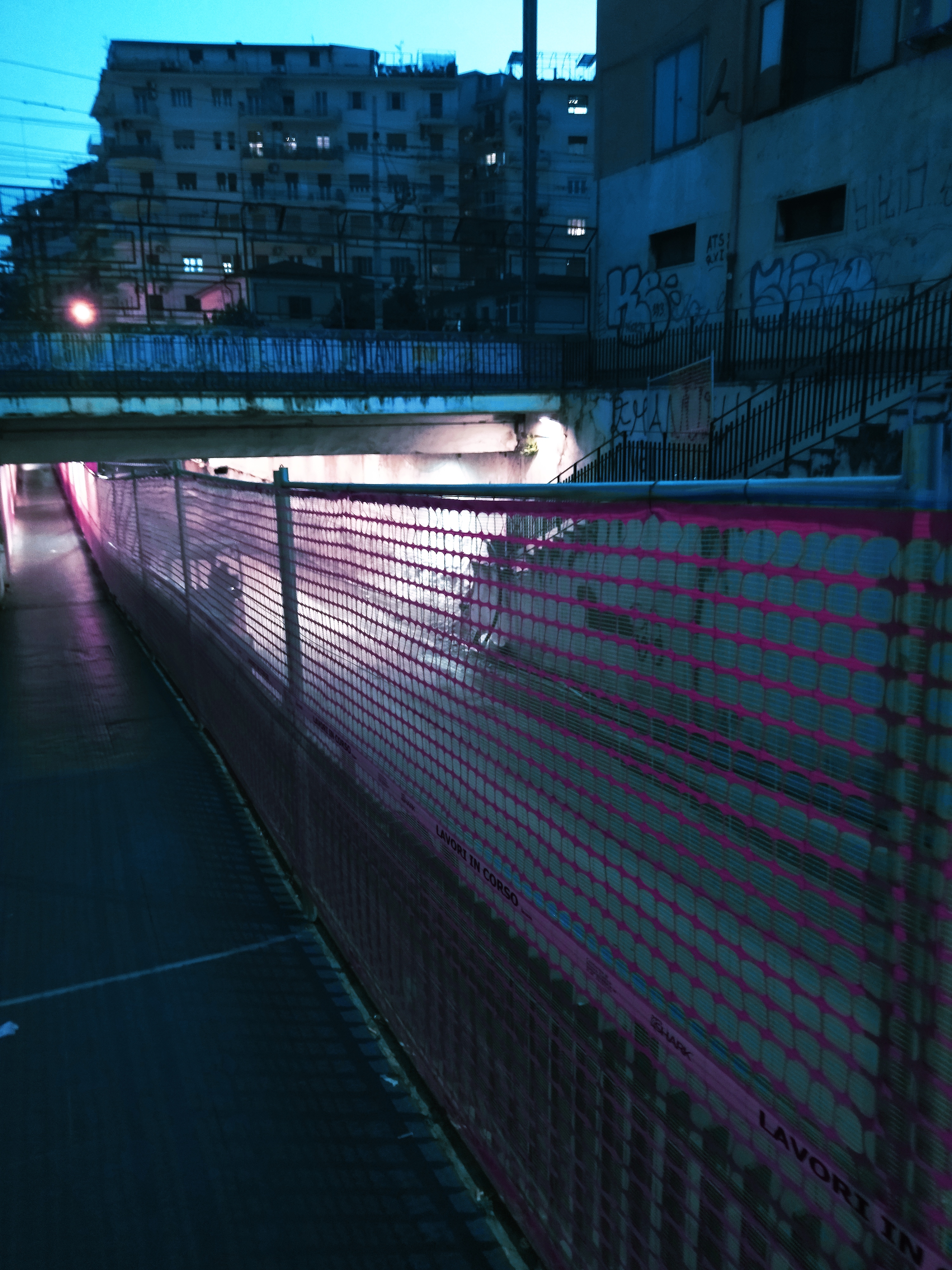 Sottopassaggio della via di ritorno verso la stazione, vista laterale di prima sera sul cantiere adiacente, filtro di luce Cyberpunk.